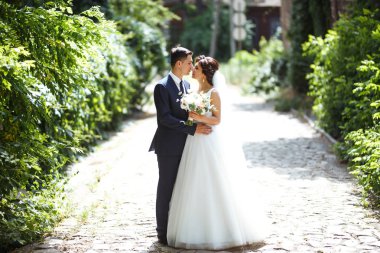 Gelin ve damat parkta birlikte yürürler. Beyaz elbiseli çekici gelin, damat koyu renkli şık bir takım elbise giyiyor. Güzel gelin ve şık damat. Düğün günü. Evlilik..