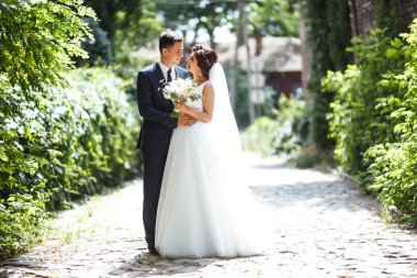 Gelin ve damat parkta birlikte yürürler. Beyaz elbiseli çekici gelin, damat koyu renkli şık bir takım elbise giyiyor. Güzel gelin ve şık damat. Düğün günü. Evlilik..