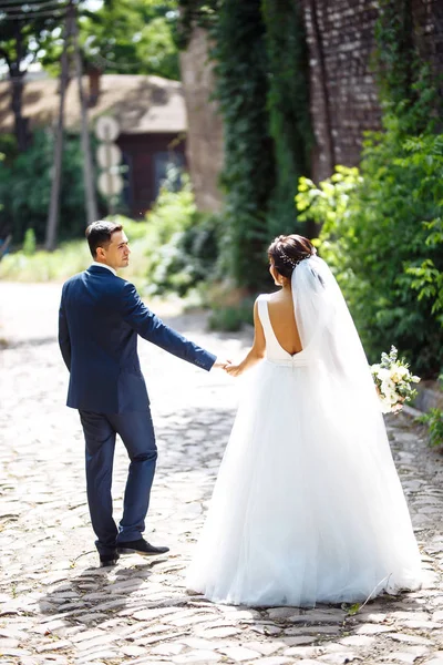 The bride and groom walk together in the park. Charming bride in a white dress, the groom is dressed in a dark elegant suit. Pretty bride and stylish groom. Wedding day. Marriage.