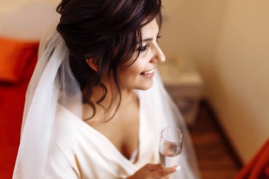 Beyaz bornozlu güzel gelin elinde bir bardak şampanya tutuyor. Profesyonel makyajlı ve saç stili olan zarif bir kadın. Gelinin düğün sabahı. Düğün günü. Gelinlik. Moda Gelini.