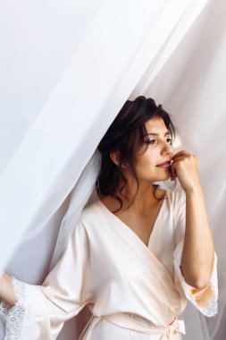 Beyaz cüppeli zarif gelin perdenin altında poz veriyor. Sabah hazırlıklarında harika bir gelin. Profesyonel makyajlı ve saç stili olan güzel bir kadın. Gelinin sabahı. Düğün günü. Moda Gelini.