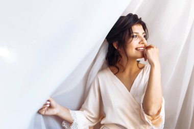 Beyaz cüppeli zarif gelin perdenin altında poz veriyor. Sabah hazırlıklarında harika bir gelin. Profesyonel makyajlı ve saç stili olan güzel bir kadın. Gelinin sabahı. Düğün günü. Moda Gelini.