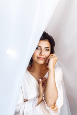 Beyaz cüppeli zarif gelin perdenin altında poz veriyor. Sabah hazırlıklarında harika bir gelin. Profesyonel makyajlı ve saç stili olan güzel bir kadın. Gelinin sabahı. Düğün günü. Moda Gelini.