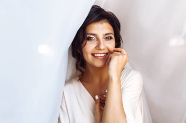 Beyaz cüppeli zarif gelin perdenin altında poz veriyor. Sabah hazırlıklarında harika bir gelin. Profesyonel makyajlı ve saç stili olan güzel bir kadın. Gelinin sabahı. Düğün günü. Moda Gelini.