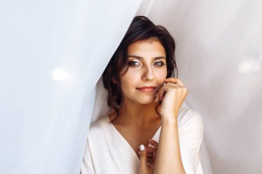 Beyaz cüppeli zarif gelin perdenin altında poz veriyor. Sabah hazırlıklarında harika bir gelin. Profesyonel makyajlı ve saç stili olan güzel bir kadın. Gelinin sabahı. Düğün günü. Moda Gelini.