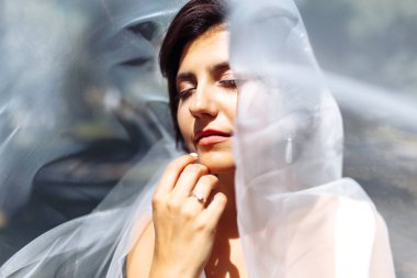 Duvağın altındaki gelinin güzel bir portresi. Profesyonel makyajlı ve saç stili olan zarif bir kadın. Bir duvağın altına gizlenmiş narin gelinlerin ellerinin fotoğrafı. Düğün günü. Evlilikten. Moda Gelini.