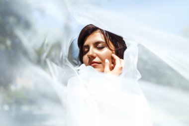 Duvağın altındaki gelinin güzel bir portresi. Profesyonel makyajlı ve saç stili olan zarif bir kadın. Bir duvağın altına gizlenmiş narin gelinlerin ellerinin fotoğrafı. Düğün günü. Evlilikten. Moda Gelini.