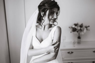 Gelinin sabahı. Beyaz elbiseli, elinde düğün buketi olan güzel, gülümseyen esmer gelin. Profesyonel makyajlı ve saç stili olan zarif bir kadın. Gelinin düğün sabahı. 