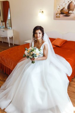 Gelinin sabahı. Beyaz elbiseli, elinde düğün buketi olan güzel, gülümseyen esmer gelin. Profesyonel makyajlı ve saç stili olan zarif bir kadın. Gelinin düğün sabahı. 