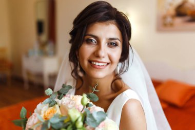 Gelinin sabahı. Beyaz elbiseli, elinde düğün buketi olan güzel, gülümseyen esmer gelin. Profesyonel makyajlı ve saç stili olan zarif bir kadın. Gelinin düğün sabahı. 