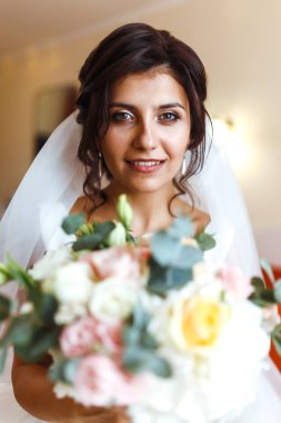 Gelinin sabahı. Beyaz elbiseli, elinde düğün buketi olan güzel, gülümseyen esmer gelin. Profesyonel makyajlı ve saç stili olan zarif bir kadın. Gelinin düğün sabahı. 