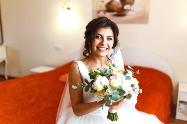 Gelinin sabahı. Beyaz elbiseli, elinde düğün buketi olan güzel, gülümseyen esmer gelin. Profesyonel makyajlı ve saç stili olan zarif bir kadın. Gelinin düğün sabahı. 