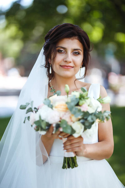 Beautiful bride with a wedding bouquet for a walk in the park. Young bride in white dress. Beautiful woman with professional make up and hair style. A beautiful bride portrait. Wedding day. Marriage.