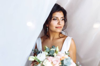 Perdenin altında çiçek demeti olan beyaz elbiseli genç gelin. Profesyonel makyajlı ve saç stili olan güzel bir kadın. Gelinin sabahı. Düğün günü. Moda Gelini.