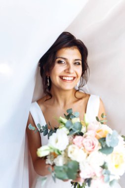 Perdenin altında çiçek demeti olan beyaz elbiseli genç gelin. Profesyonel makyajlı ve saç stili olan güzel bir kadın. Gelinin sabahı. Düğün günü. Moda Gelini.