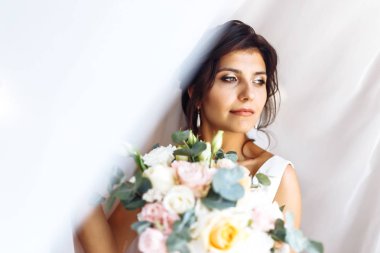 Perdenin altında çiçek demeti olan beyaz elbiseli genç gelin. Profesyonel makyajlı ve saç stili olan güzel bir kadın. Gelinin sabahı. Düğün günü. Moda Gelini.