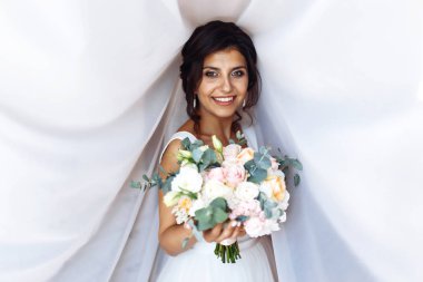 Perdenin altında çiçek demeti olan beyaz elbiseli genç gelin. Profesyonel makyajlı ve saç stili olan güzel bir kadın. Gelinin sabahı. Düğün günü. Moda Gelini.