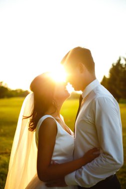 Gün batımında harika bir çift. Gelin ve damat bir buket çiçekle birlikte gün batımında yeşil tarlanın arka planına karşı ellerinde olacaklar. Romantik Evli Genç Aile. 