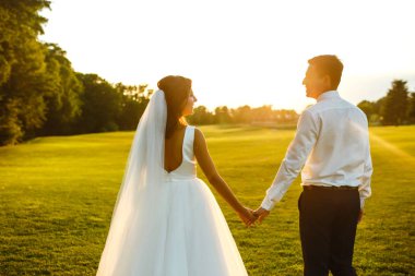 Gün batımında harika bir çift. Gelin ve damat bir buket çiçekle birlikte gün batımında yeşil tarlanın arka planına karşı ellerinde olacaklar. Romantik Evli Genç Aile. 