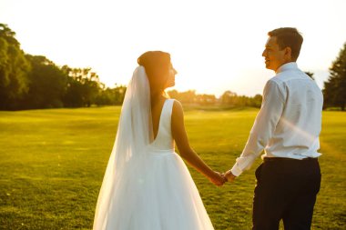 Gün batımında harika bir çift. Gelin ve damat bir buket çiçekle birlikte gün batımında yeşil tarlanın arka planına karşı ellerinde olacaklar. Romantik Evli Genç Aile. 