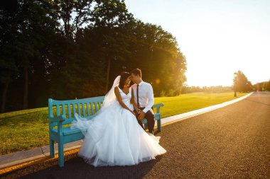 Mutlu bir gelin ve damat gün batımında bir bankta birlikte oturuyorlar. Aşıklar parkta bir bankta sarılır. Uzun beyaz elbiseli güzel gelin. Genç bir aile. Evlilikten. Düğün konsepti.