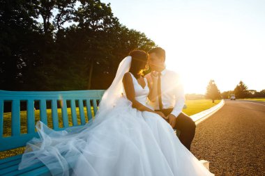 Mutlu bir gelin ve damat gün batımında bir bankta birlikte oturuyorlar. Aşıklar parkta bir bankta sarılır. Uzun beyaz elbiseli güzel gelin. Genç bir aile. Evlilikten. Düğün konsepti.