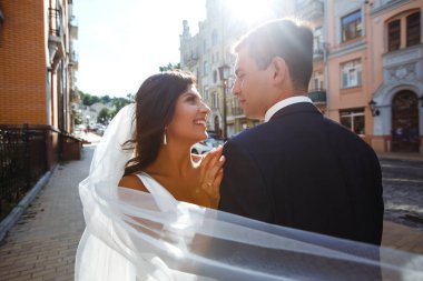 Gelin ve damat, uçan bir duvağın gölgesinde nazikçe öpüşürler. Siyah takım elbiseli şık bir damat uçan peçeli beyaz elbiseli güzel bir gelini nazikçe kucaklıyor. Birlikte. Düğün için. Aşk.