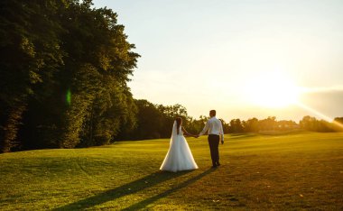Mutlu gelin ve damat gün batımına gidiyor. Gün batımında harika bir çift. Gelin ve damat, gün batımında yeşil tarlanın arka planına karşı ellerinde olacak. Birlikte. Düğün.. 