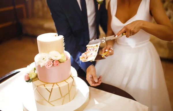 Happy bride and a groom is cutting their stylish wedding cake on wedding banquet. Newlyweds holding knife and cutting together wedding cake decorated with flowers. Sweet cake for couple.