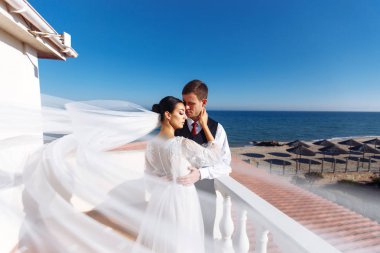 Yeni evliler, güzel bir deniz manzarasının arka planındaki uçan bir duvağın gölgesinde şefkatle eğleniyorlar. Gelin ve damat denizin arka planında sarılıp öpüşüyorlar.. 