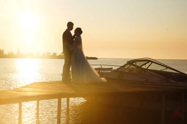 Güzel gelin ve şık damat gün batımında teknenin arka planına karşı köprüde yan yana. Yeni evliler gün batımında şefkatle sarılır, öpüşür ve eğlenirler. Düğün için. Sevgi. Romantik an. 