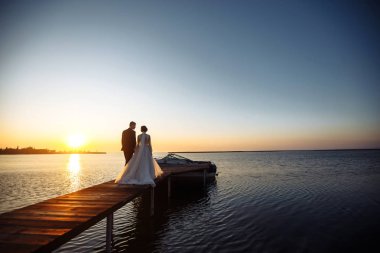 Güzel gelin ve şık damat gün batımında teknenin arka planına karşı köprüde yan yana. Yeni evliler gün batımında şefkatle sarılır, öpüşür ve eğlenirler. Düğün için. Sevgi. Romantik an. 