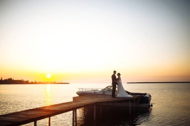 Güzel gelin ve şık damat gün batımında teknenin arka planına karşı köprüde yan yana. Yeni evliler gün batımında şefkatle sarılır, öpüşür ve eğlenirler. Düğün için. Sevgi. Romantik an. 