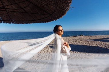 Güneşin altında beyaz elbiseli güzel gelin kumlu sahilde yürüyor. Lüks makyajlı ve saç stili olan mutlu gelin. Güneş ve mavi gökyüzünde zarif bir gelin. Düğün günü..