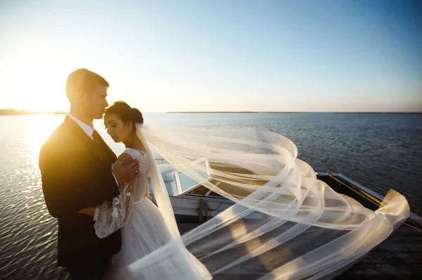 Pretty bride and stylish groom together on the bridge against the background of the boat. Newlyweds enjoy each other tenderly in the shadow of a flying veil. Together. Wedding. Love.