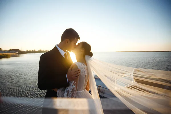 かなりの花嫁とスタイリッシュな新郎一緒に船の背景に橋の上に 新婚旅行は 空飛ぶベールの影の中で互いに優しいお楽しみください 一緒に結婚式愛 — ストック写真