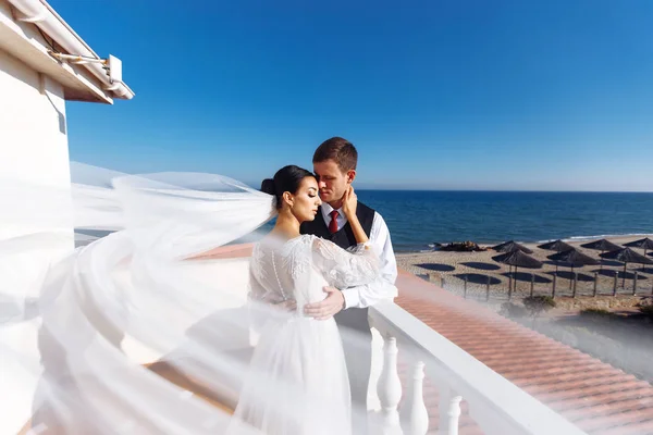 Newlyweds enjoy each other tenderly in the shadow of a flying veil on the background of a beautiful sea landscape. The bride and groom hugging and kissing on the background of the sea.