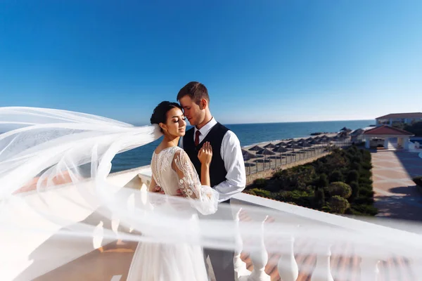 Newlyweds enjoy each other tenderly in the shadow of a flying veil on the background of a beautiful sea landscape. The bride and groom hugging and kissing on the background of the sea.