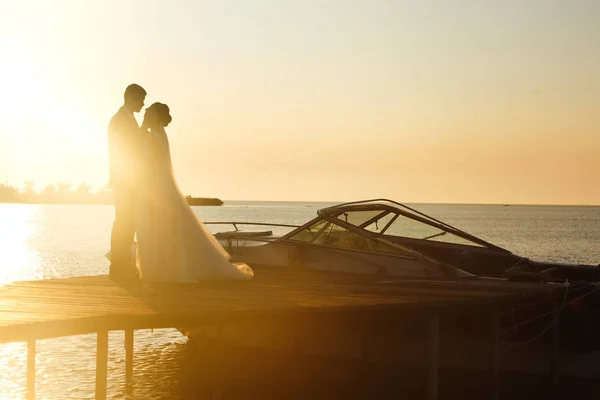夕日の船の背景に橋の上で一緒に美しい花嫁とスタイリッシュな新郎 新婚夫婦は優しく抱擁し キスをし 日没時に楽しみます 結婚式愛だロマンチックな瞬間 — ストック写真
