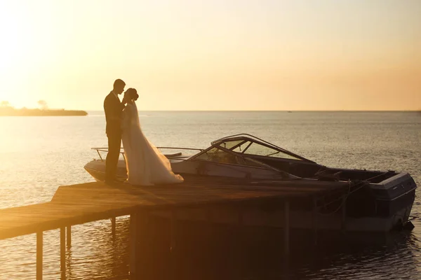 夕日の船の背景に橋の上で一緒に美しい花嫁とスタイリッシュな新郎 新婚夫婦は優しく抱擁し キスをし 日没時に楽しみます 結婚式愛だロマンチックな瞬間 — ストック写真