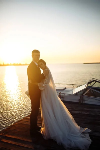 夕日の船の背景に橋の上で一緒に美しい花嫁とスタイリッシュな新郎 新婚夫婦は優しく抱擁し キスをし 日没時に楽しみます 結婚式愛だロマンチックな瞬間 — ストック写真