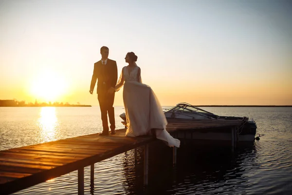 夕日の船の背景に橋の上で一緒に美しい花嫁とスタイリッシュな新郎 新婚夫婦は優しく抱擁し キスをし 日没時に楽しみます 結婚式愛だロマンチックな瞬間 — ストック写真