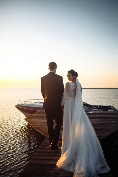 夕日の船の背景に橋の上で一緒に美しい花嫁とスタイリッシュな新郎 新婚夫婦は優しく抱擁し キスをし 日没時に楽しみます 結婚式愛だロマンチックな瞬間 — ストック写真