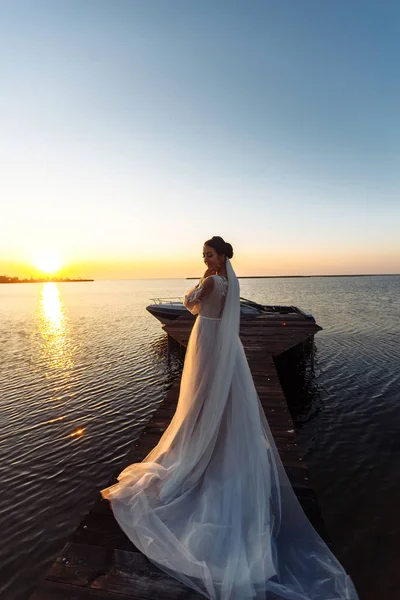 美丽的新娘 身穿雅致的白衣 靠着大海的背景 在夕阳的余晖中 站在一座桥上 拥有奢华妆容和发型的快乐新娘 婚礼日 — 图库照片