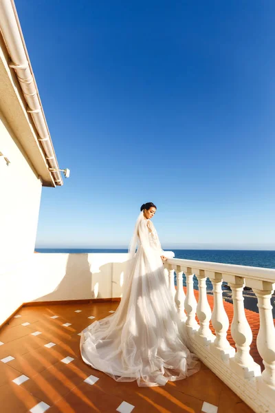 Beautiful bride with dark hair in luxurious wedding dress posing in balcony with sea view. Happy bride with luxury make-up and hairstyle. Luxurious elegant bride in a white dress in the sun.