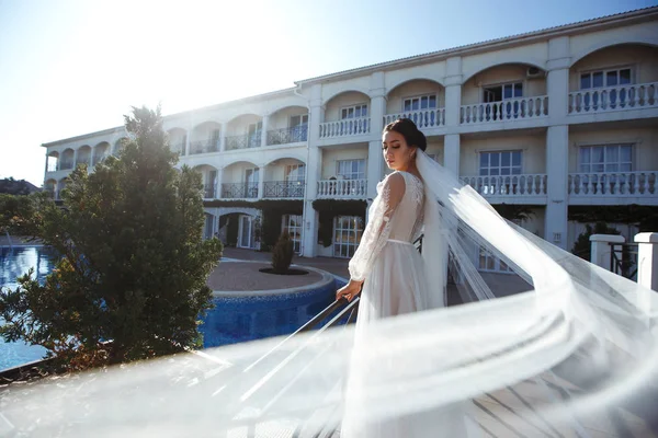 Beautiful sexy bride in white dress posing  near the pool. Happy bride  in  luxurious wedding dress posing in the sunshine. Wind blows her veil. Wedding day. Marriage. Fashion bride.