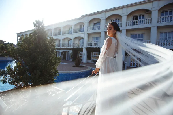 Beautiful sexy bride in white dress posing  near the pool. Happy bride  in  luxurious wedding dress posing in the sunshine. Wind blows her veil. Wedding day. Marriage. Fashion bride.