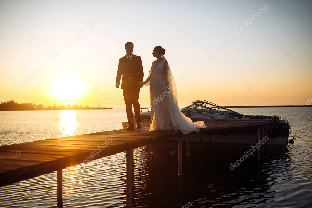 Beautiful bride and stylish groom together on the bridge against the background of the boat at sunset. Newlyweds tenderly hug, kiss and enjoy each other at sunset. Wedding. Love. Romantic moment. 