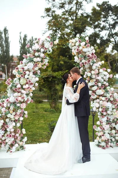 Loving newlyweds stand near the flower arch. Young beautiful happy wedding couple is kissing near arch at the ceremony. Pretty bride and stylish groom. Wedding and honeymoon concept. Love.