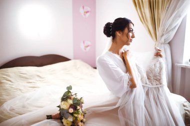 Gelinliğin yanındaki güzel gelinin sabahı. Beyaz cüppeli gelin gelinliğini tutuyor ve ona hayran. Profesyonel makyajlı ve saç stili olan güzel bir kadın. Gelinin sabahı. 